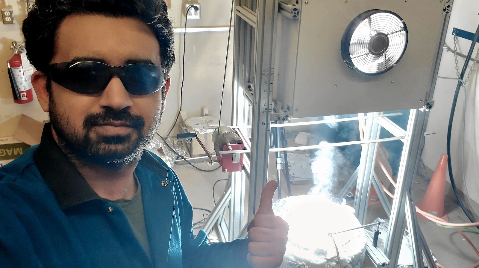 Researcher standing in front of equipment with bright lights. Researcher wearing PPE and sunglasses.