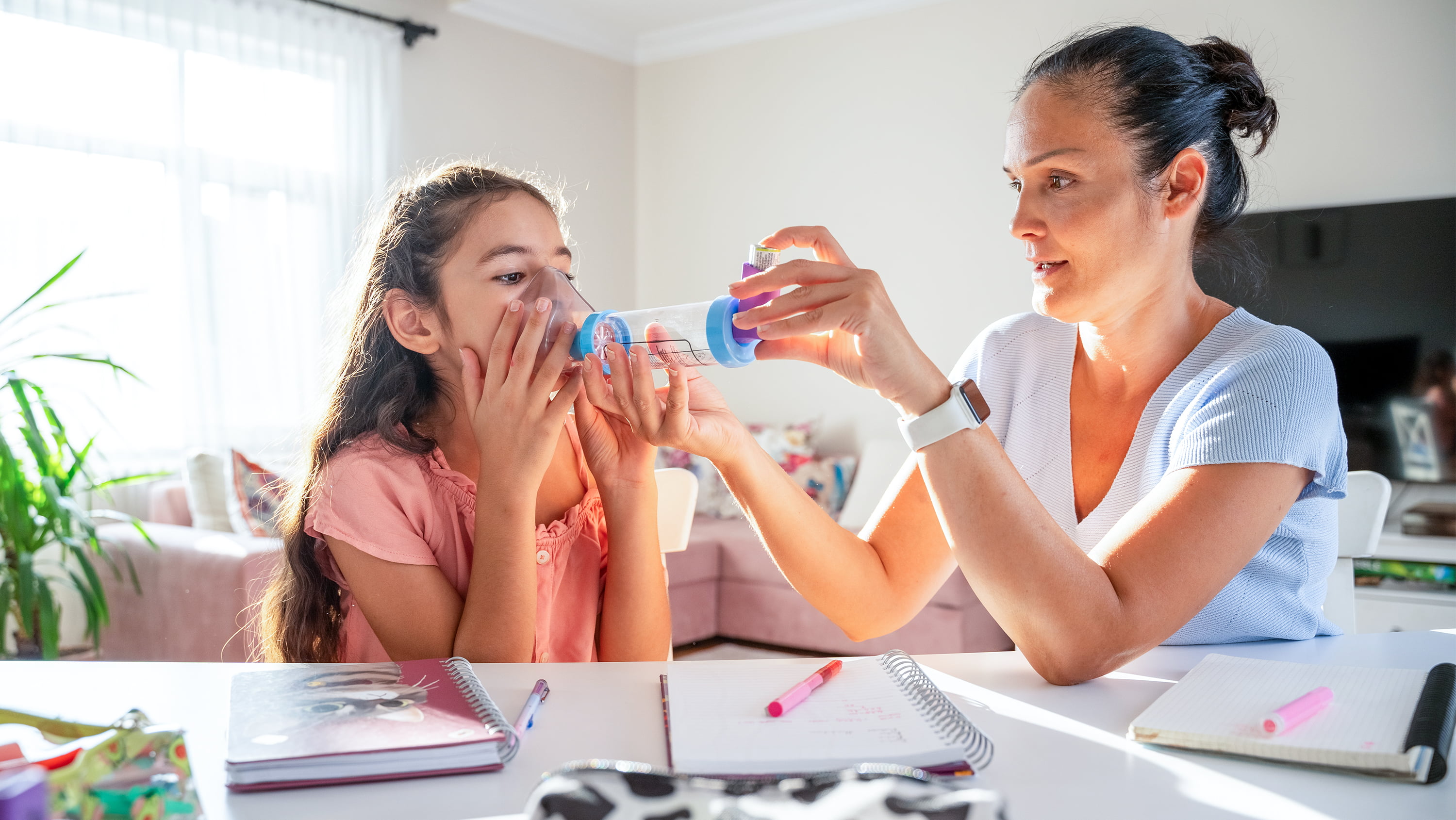 A mother administers an ashthma inhaler to her daughter. Both are seated at a table littered with notebooks. (Photo: Getty Images)