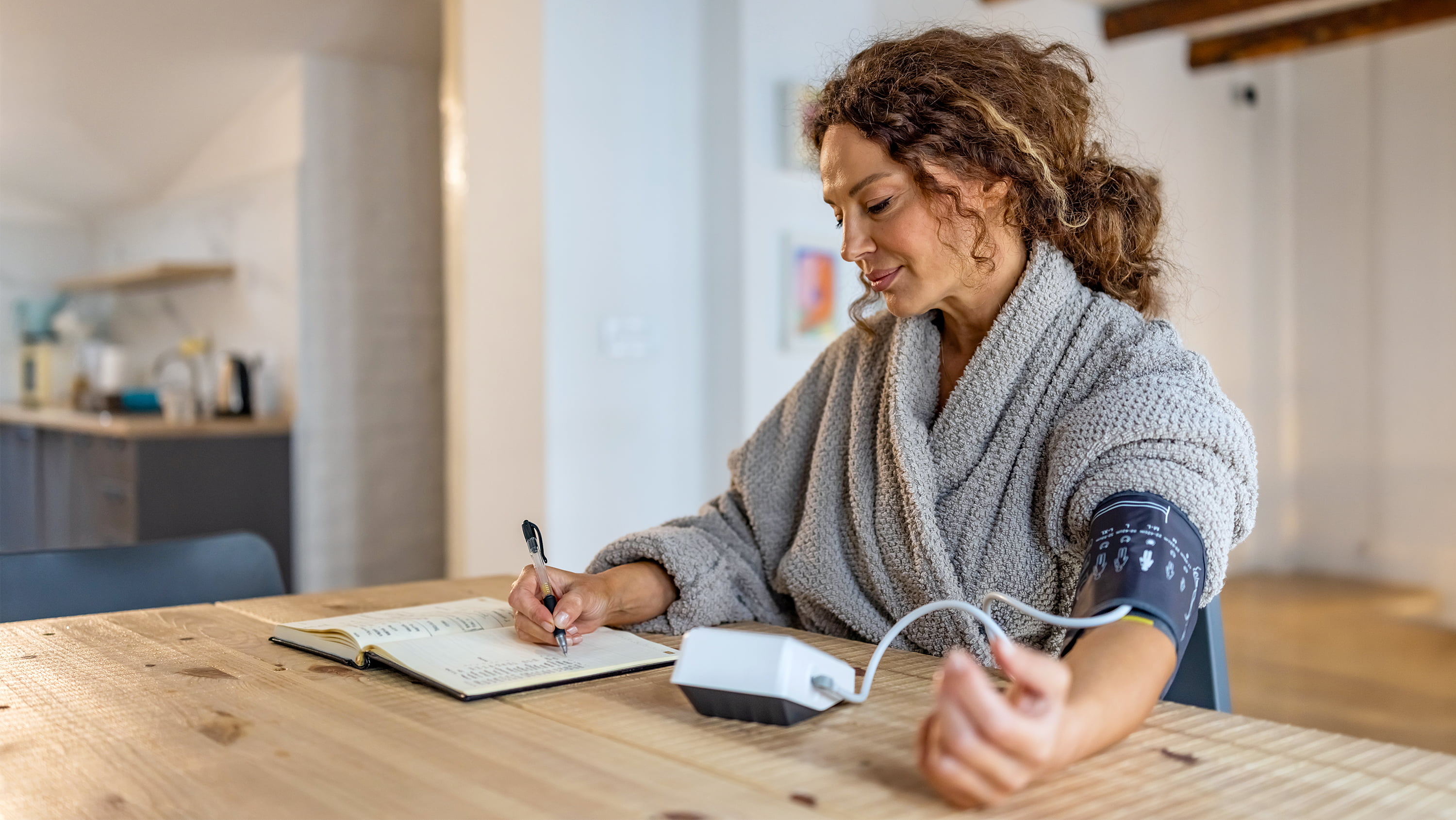 A woman seated at a table smiles softly as she takes her blood pressure and records the results in a notebook. (Photo: Getty Images)