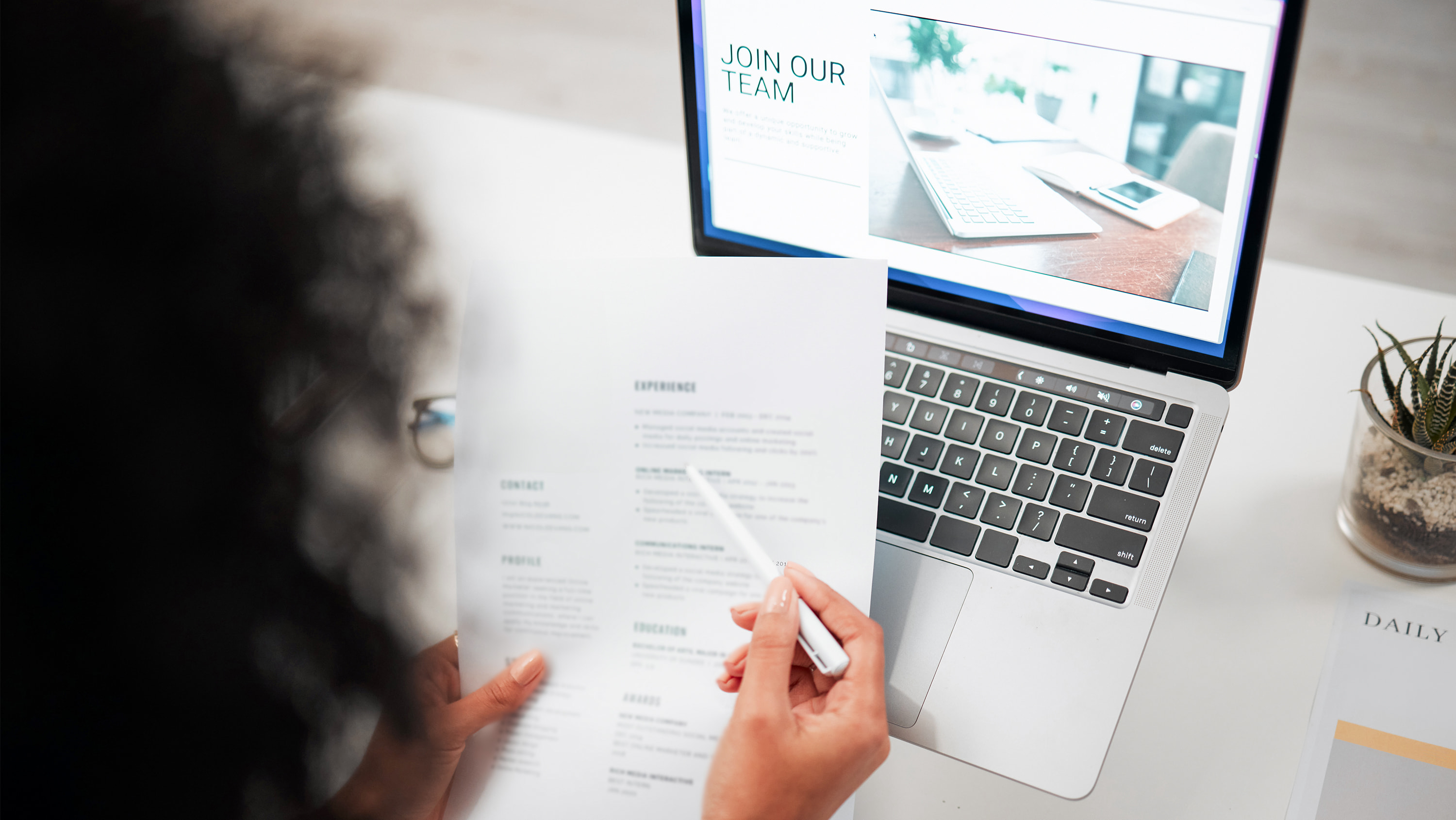 An over-the-shoulder view shows a person holding a printed resume and pen while sitting in front of an open laptop displaying a "JOIN OUR TEAM" webpage. (Photo: Getty Images)