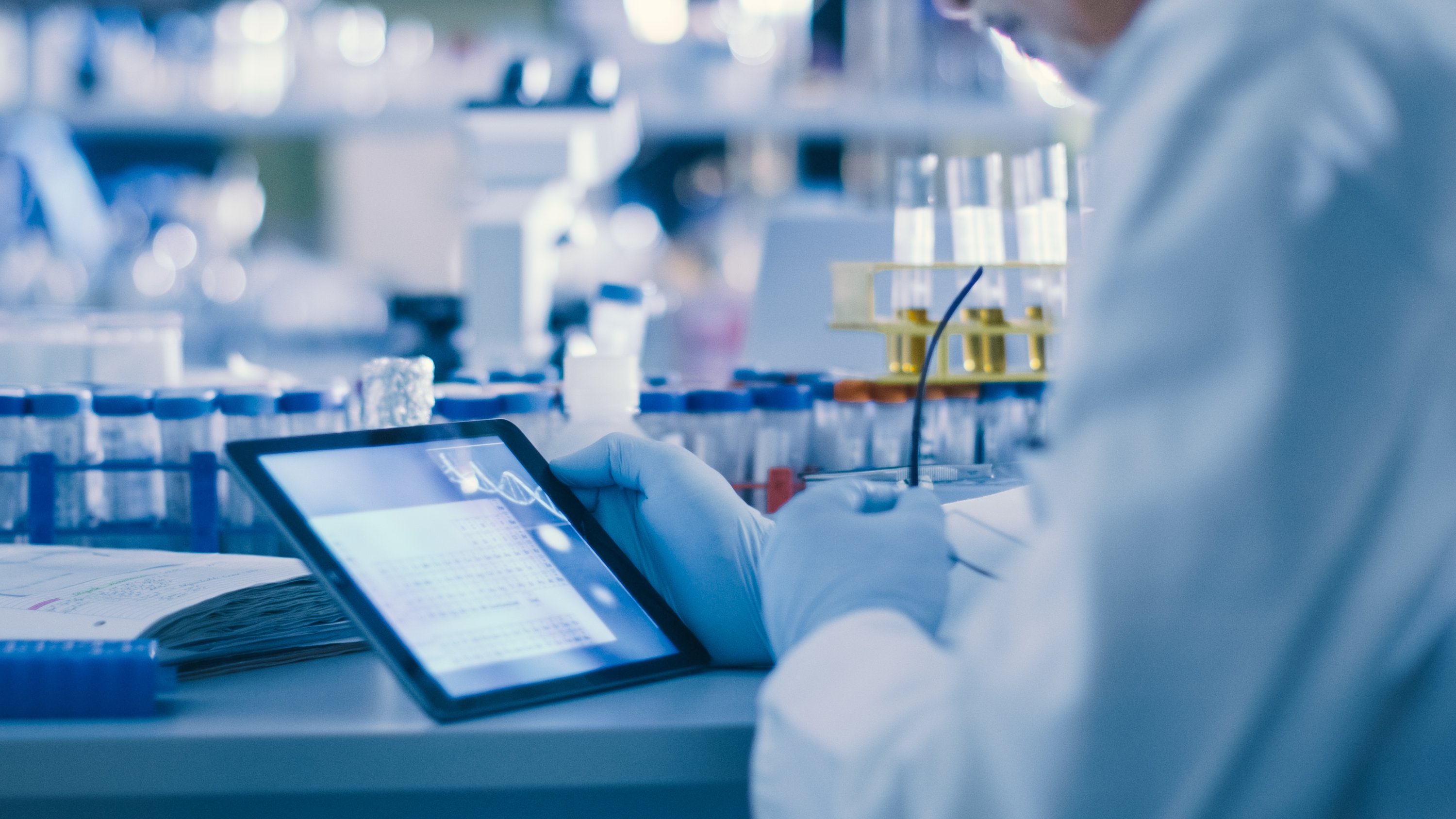 A scientist wearing blue gloves holds a tablet displaying genetic data while taking notes in a laboratory filled with test tubes and scientific equipment. (Photo: Getty Images)
