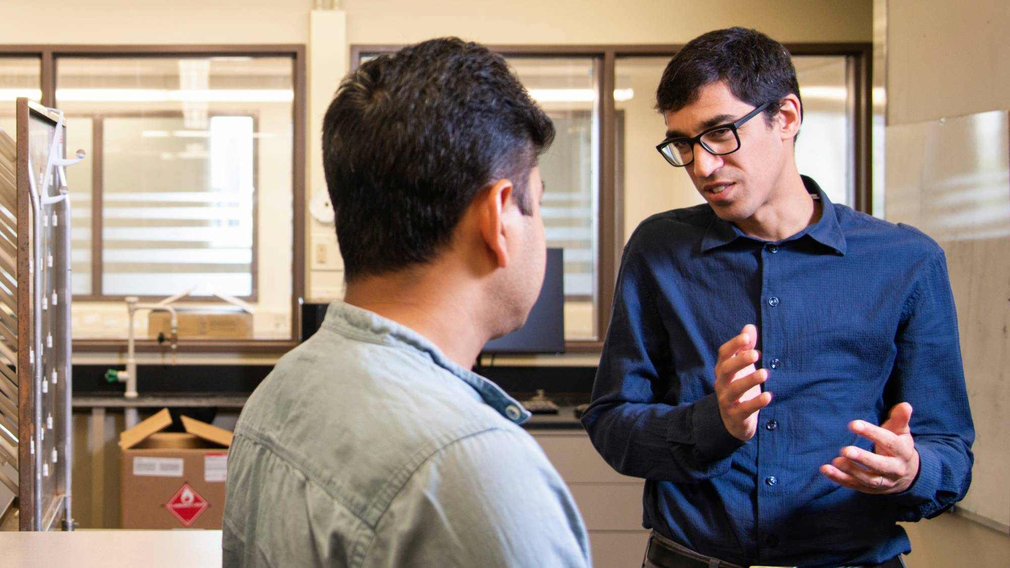 Two people have a conversation in a laboratory, with one person gesturing while speaking and the other listening.
