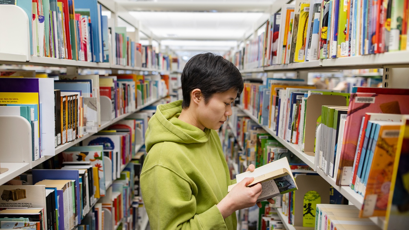 A student wearing a light green hoodie stands between library bookshelves, looking down and reading a book.