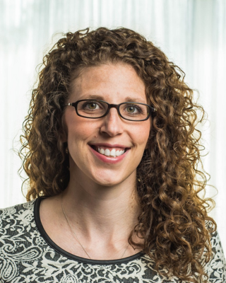 A person with long curly hair and glasses wearing a patterned top smiles at the camera