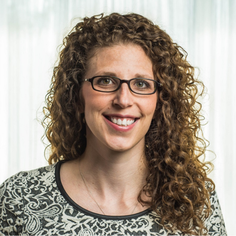 A person with long curly hair and glasses wearing a patterned top smiles at the camera