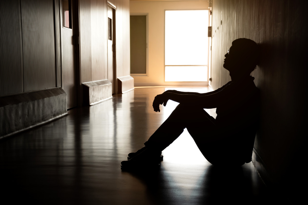 A person sits alone in a dark, empty hallway, silhouetted by light from the end of the hall.