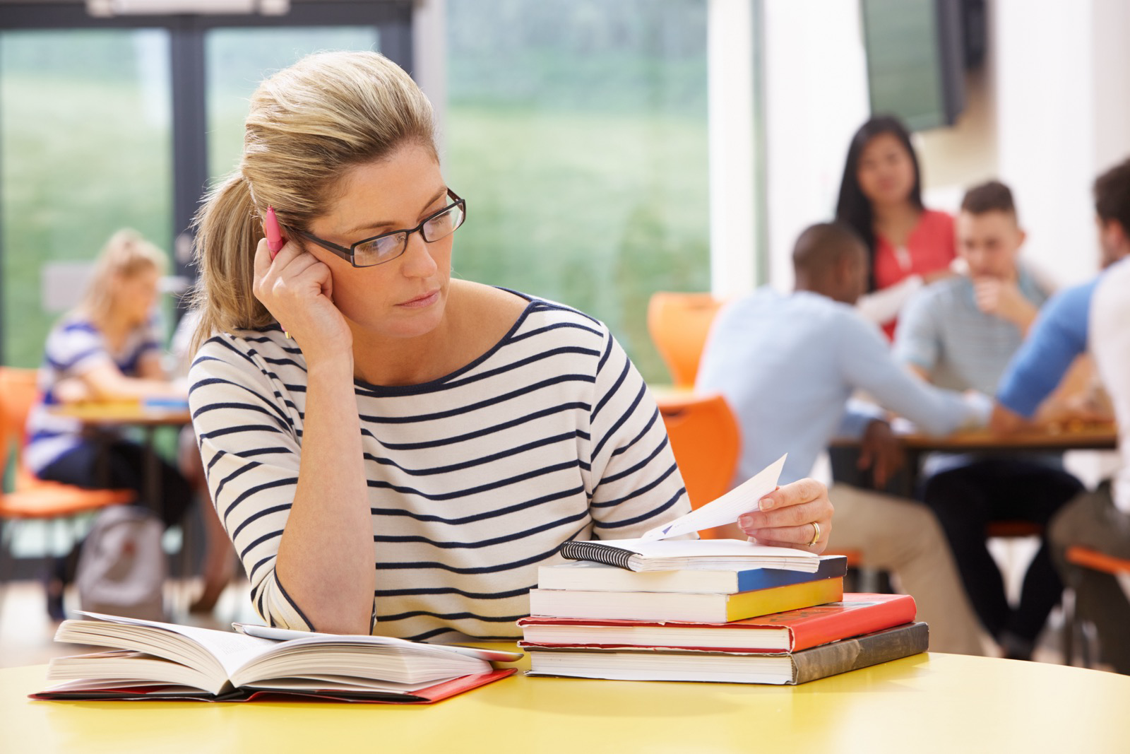 Mature female student in a classroom looking at papers and a stack of book. Other students studing and engaging at tables behind her. They are blurry.