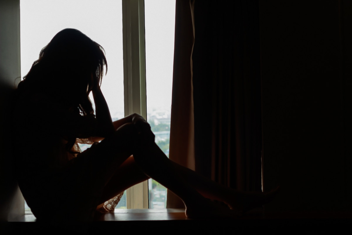 A young woman sits alone in a dark room, head in her hands.