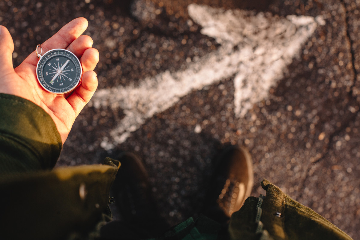 A hand holds a compass, with a person looking down towards it. At their feet, an arrow is painted on the ground.
