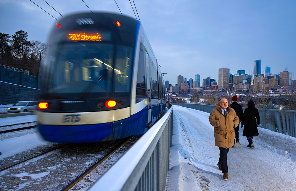 Winter photo of Murtaza Haider walking on a pedestrian pathway to the right of the train on an Edmonton LRT line, downtown Edmonton in the background.