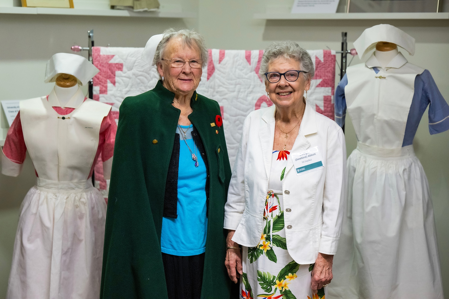 Two women posing with vintage nurse uniforms on mannequins in a museum exhibit. One woman wears a green cape over a blue top, the other a white jacket with a floral skirt.