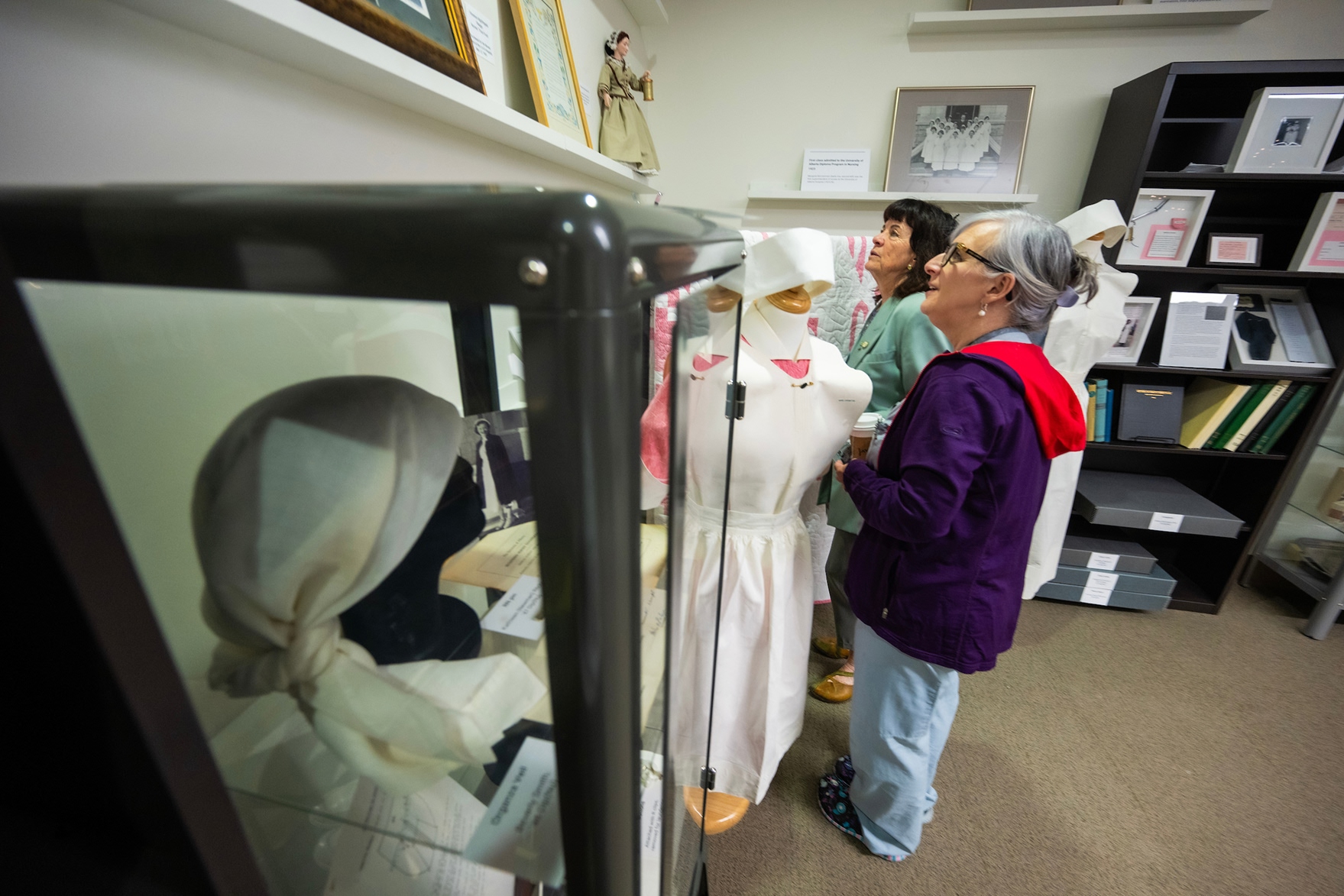 An woman in a purple jacket viewing a glass display case containing a vintage nurse uniform with a white bonnet in a museum setting.