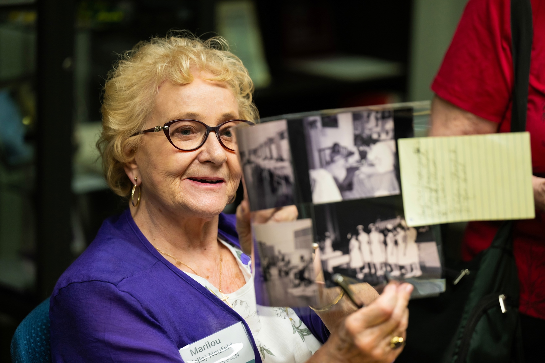 Close-up of a smiling elderly woman with glasses and a purple top holding black-and-white historical photographs and a yellow note during a museum visit.