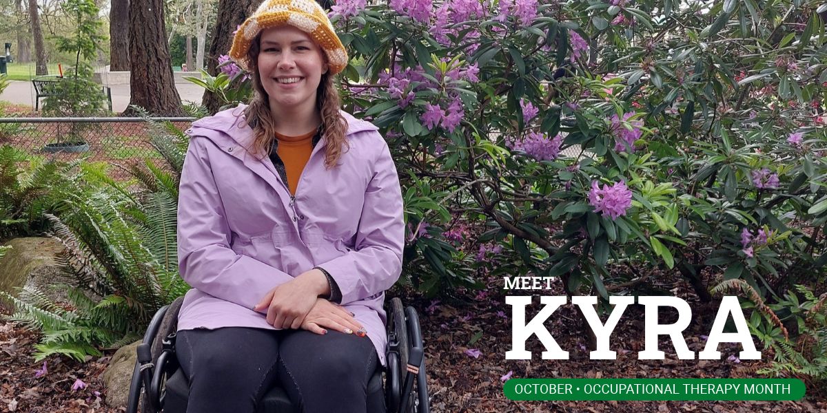 Kyra Mazer smiling in her wheelchair on a garden path, with flowering rhododendrons behind her