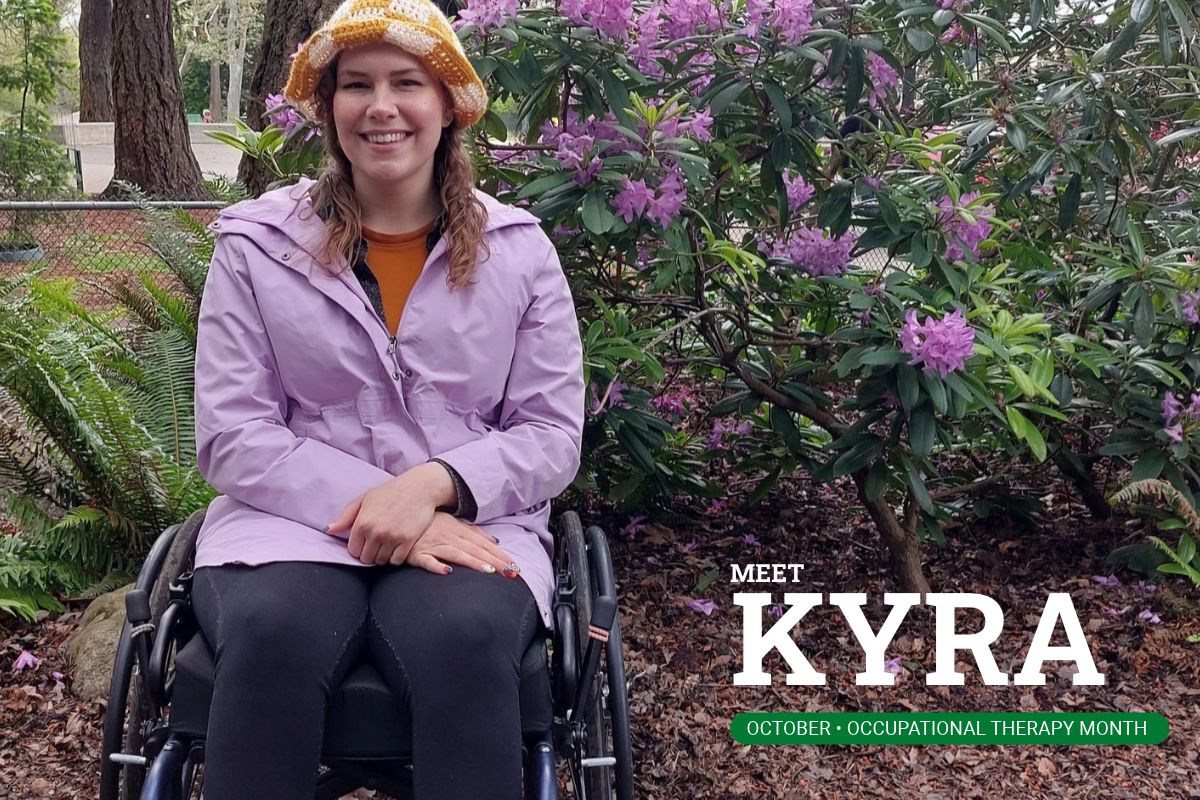 Kyra Mazer smiling in her wheelchair on a garden path, with flowering rhododendrons behind her