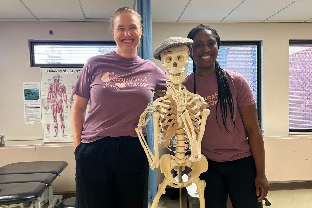Two OT students posing beside a skeleton in a clinic setting