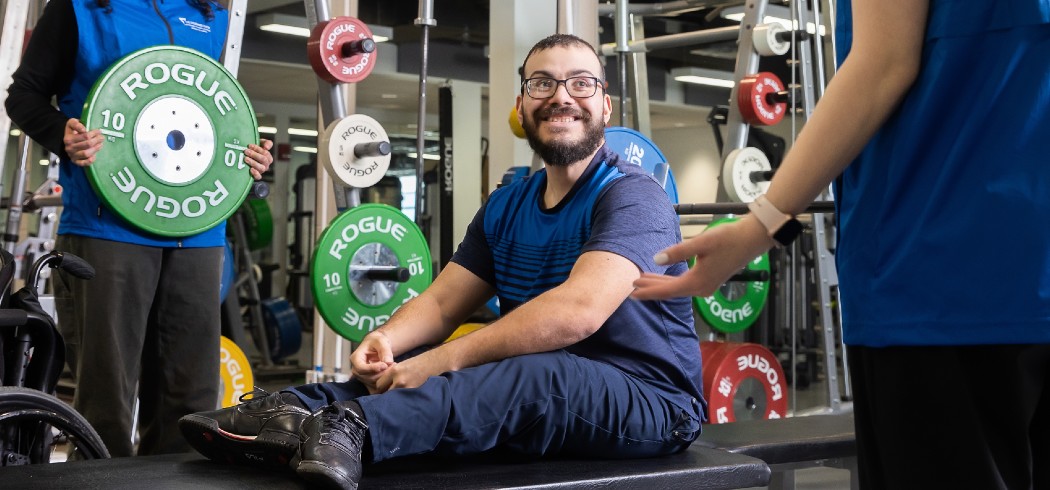Man siting on padded table with fitness instructors holding large weights beside hime
