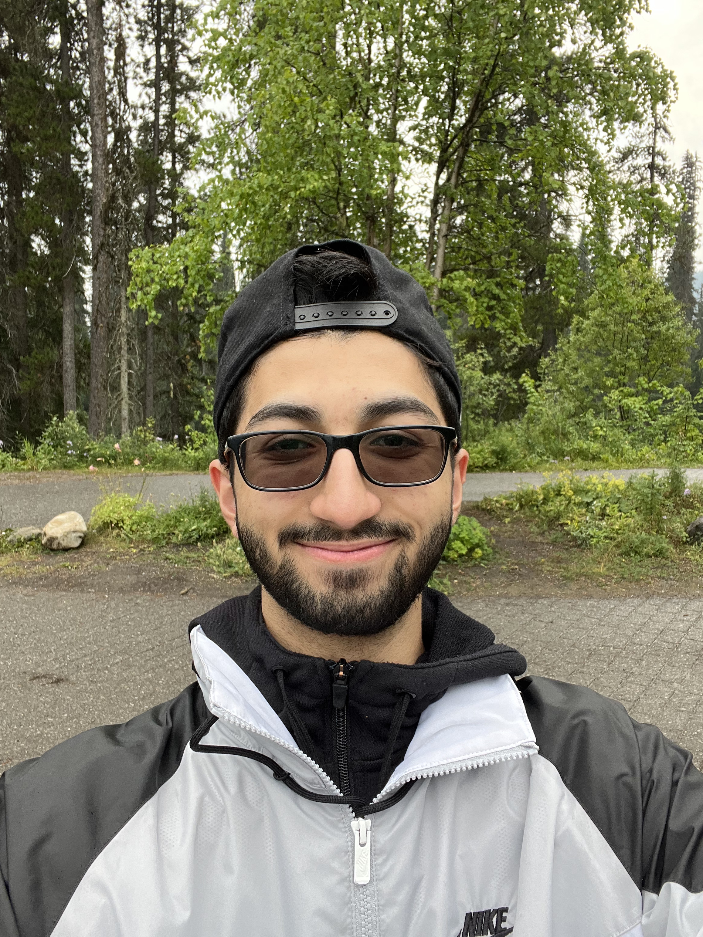 A young persian guy stands in the forest. He is wearing sunglasses and backward baseball cap