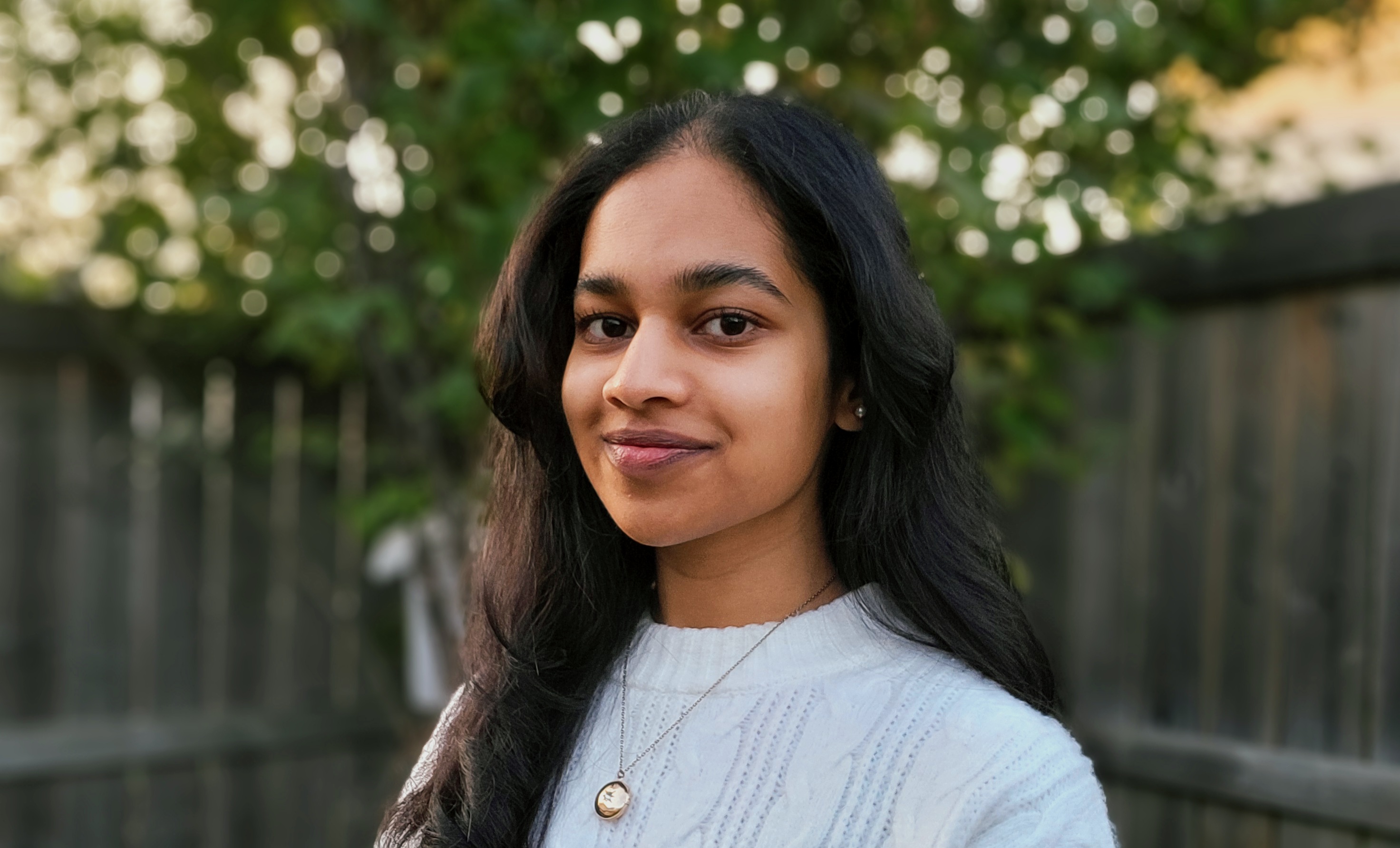 A young south asian woman stands in front of a wooden fence.