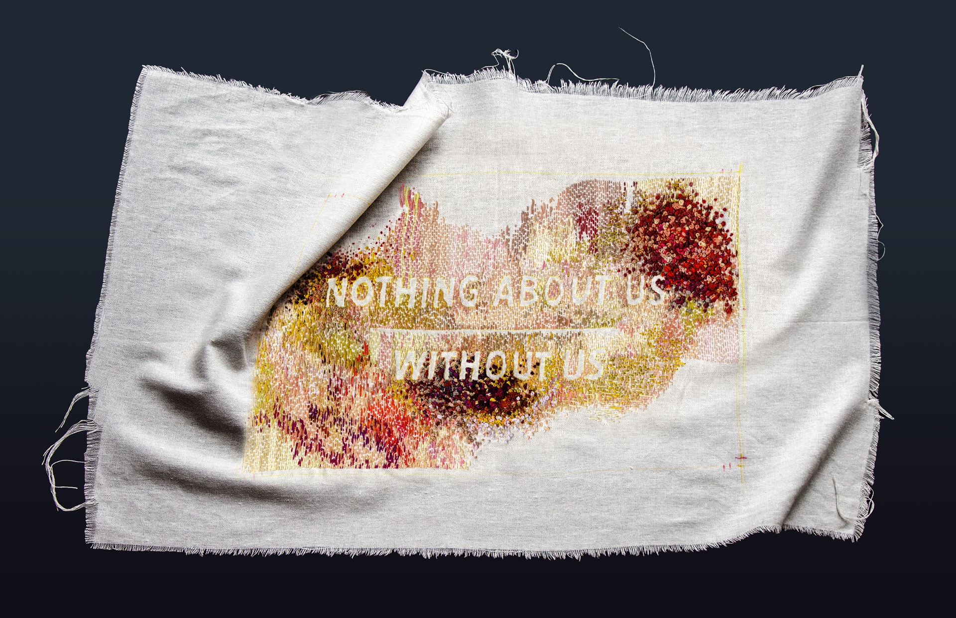 A frayed rectangle of white linen rests in a dark undefined space. At the centre of the cloth, disability justice’s prolific call “NOTHING ABOUT US WITHOUT US” rests in stark relief against colourful blooms of embroidery floss. Short vertical dashes and twisted knots in corporeal hues of reds, purples, and greenish yellows coalesce to form a palette of bruising and healing. 