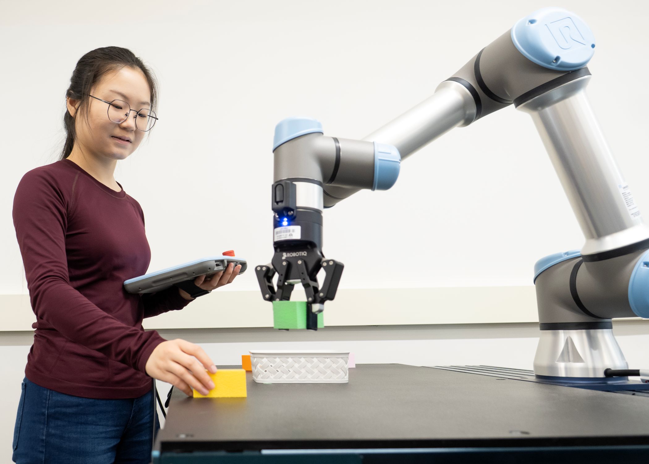 Graduate teaching assistant Yuezhen Gao operates and monitors a robotic arm performing object manipulation tasks using a tablet controller in a collaborative robotics experiment.