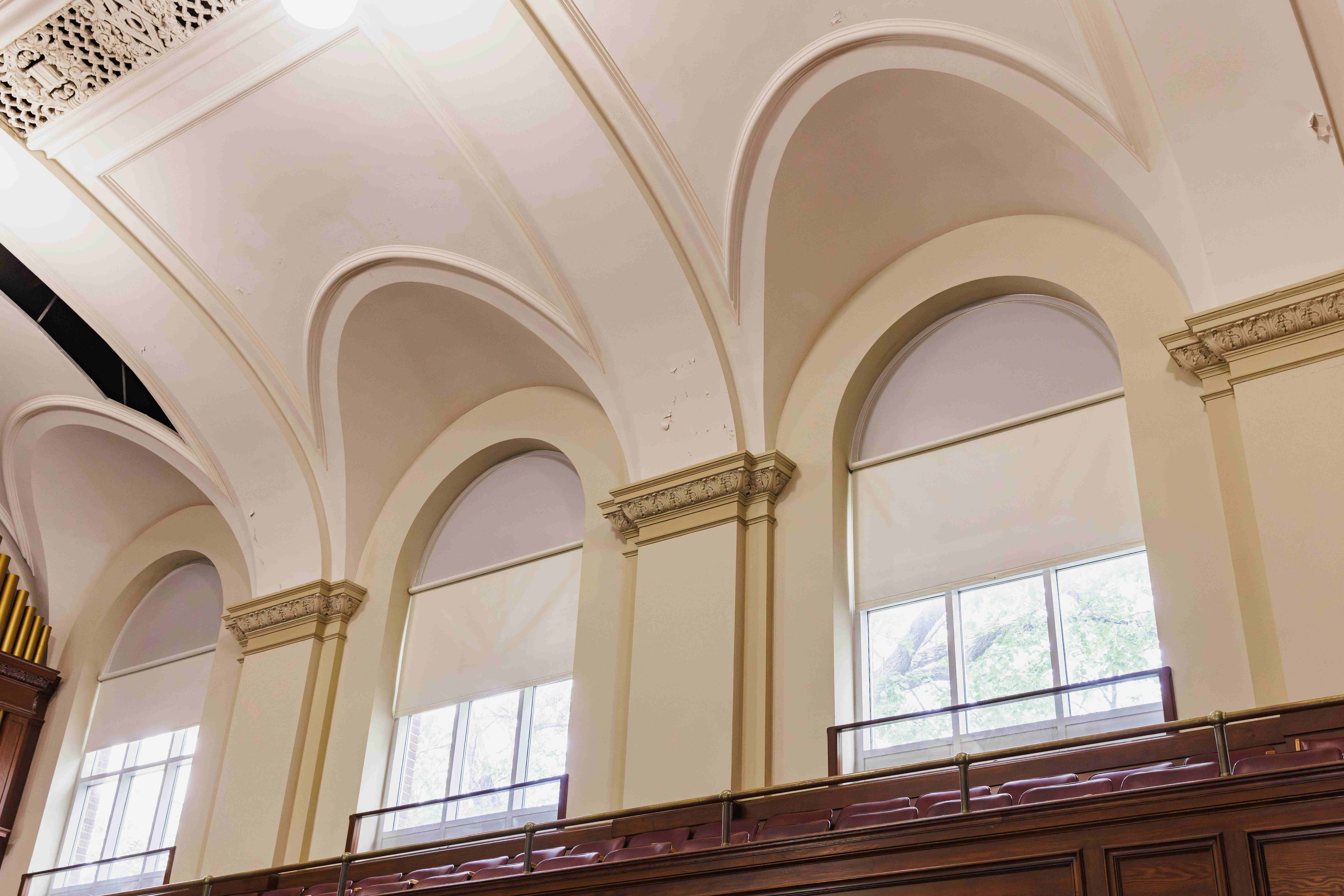 Historic interior featuring soaring vaulted ceilings, arched windows, decorative columns, and tiered seating illuminated by natural light.