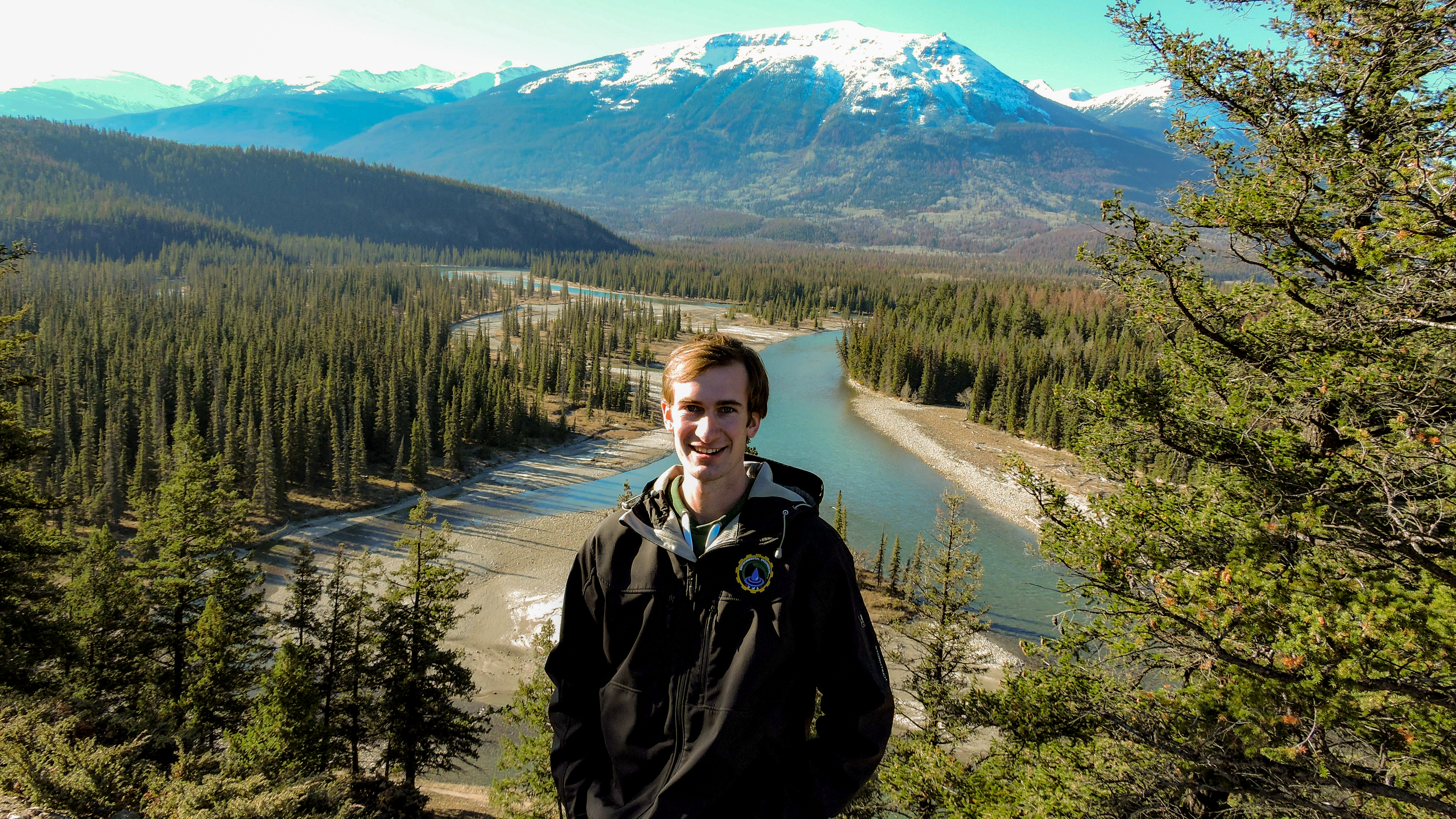 Evan stands in front of a mountain valley with trees and a stream