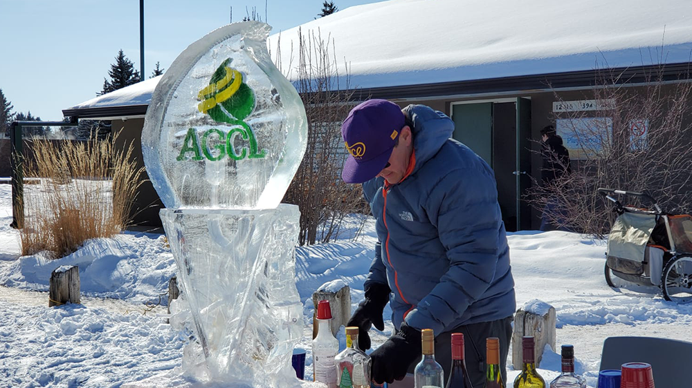 A man serves drinks at a community league function outside on a sunny winter day, next to a large ice sculpture featuring the league's logo