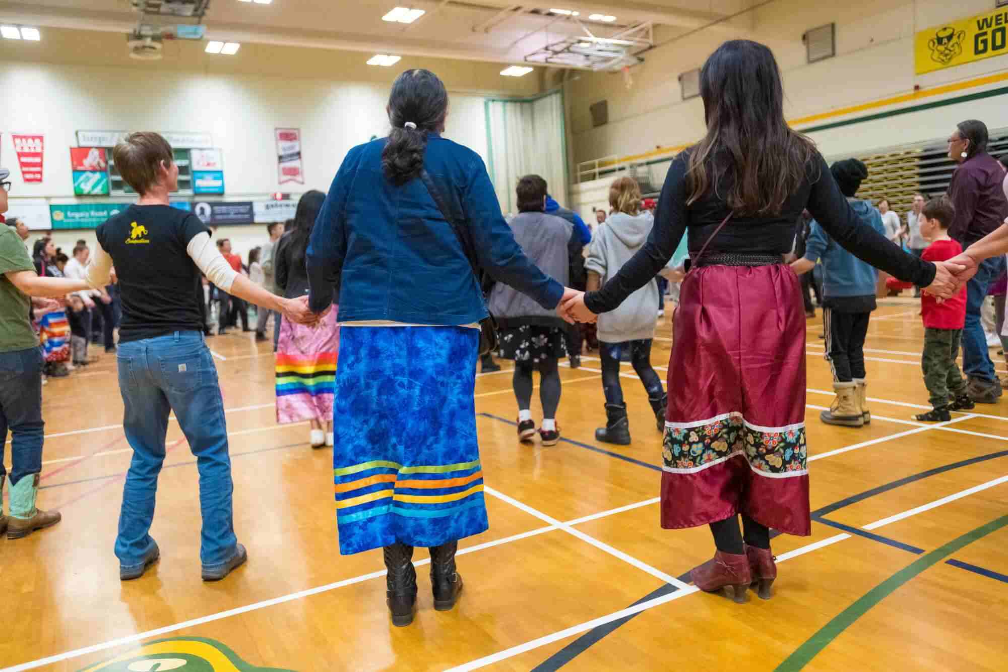 A group of people, some of them wearing ribbon skirts, holding hands at a Round Dance