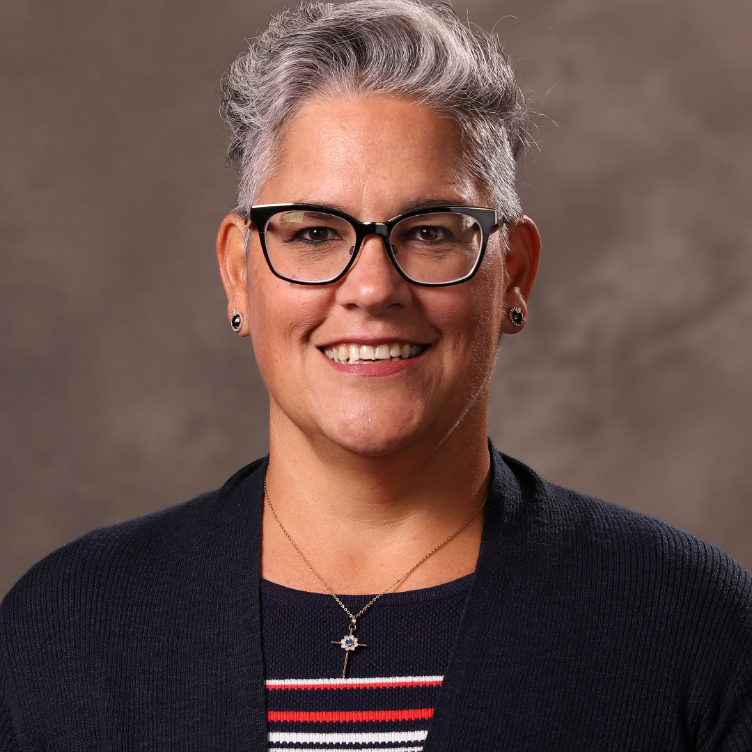Metis woman with short, grey hair wearing black-rimmed glasses and a black shirt smiles directly into camera