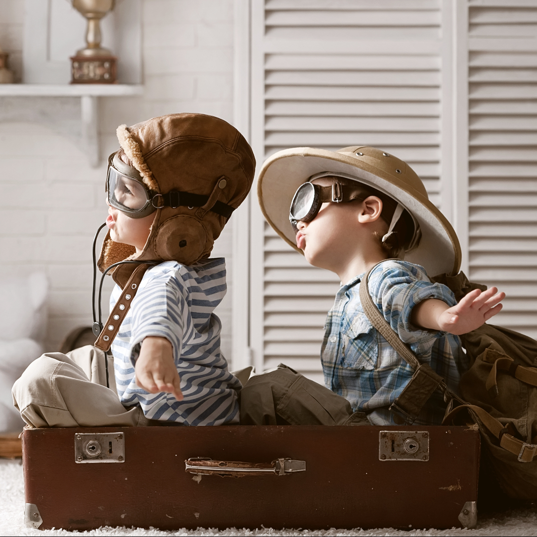 two toddler-aged males play make-believe in a airplane made out of boxes, wearing aviator hats and googles, sitting in what appears to be a living room 