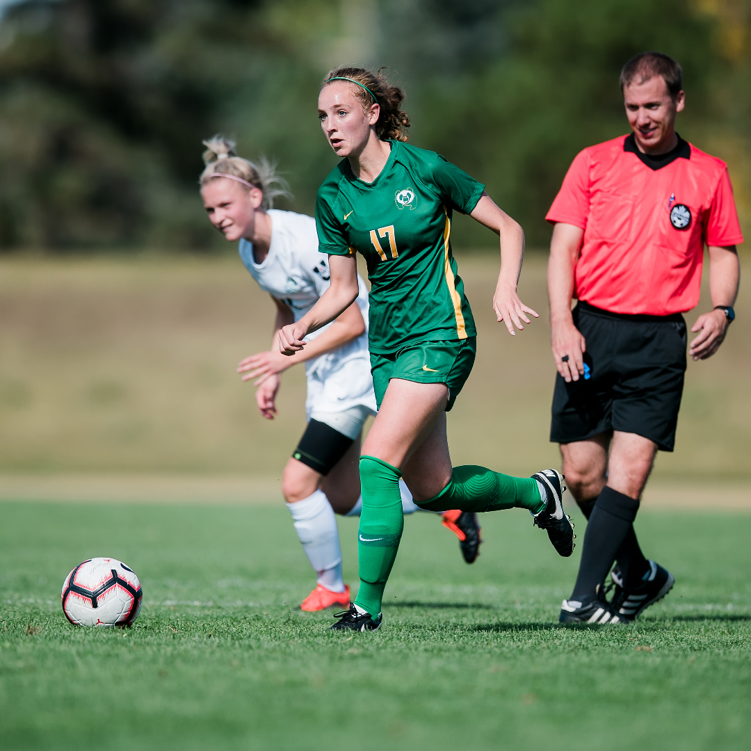 female soccer player in all green uniform ball handles up the field with an opponent and referee trailing behind her