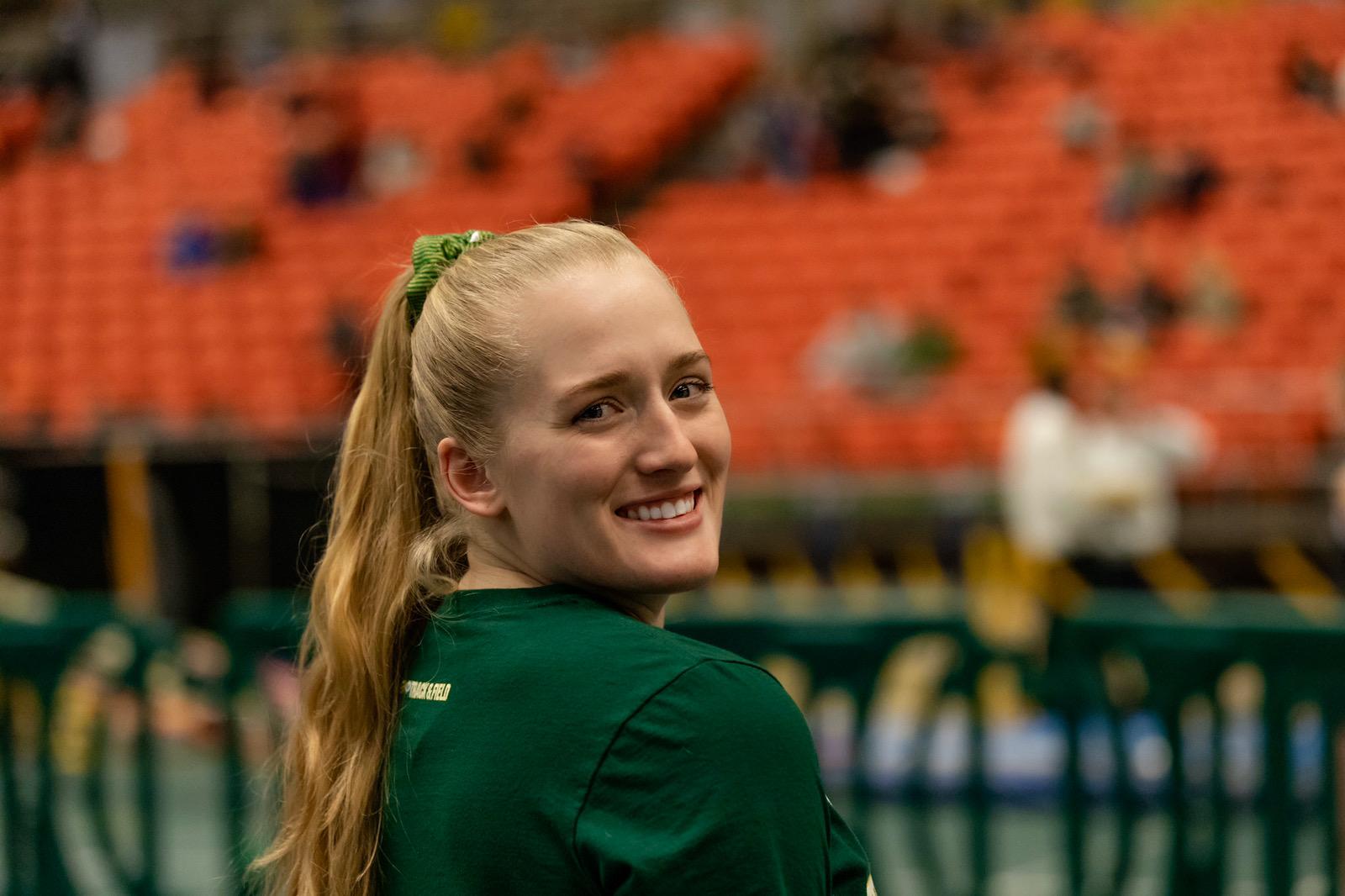 young woman looking over her right shoulder, smiling into the camera in a gymnasium with bleacher blurred out in background