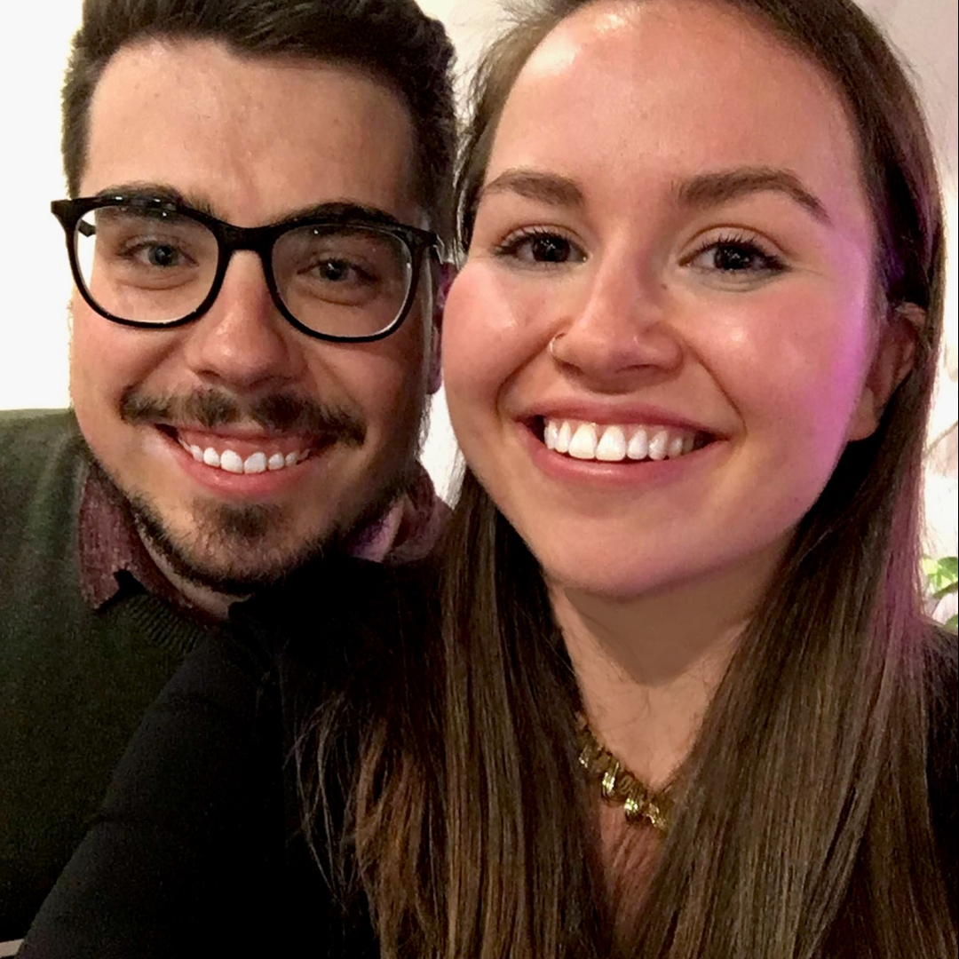 brunette male with glasses and facial hair in green sweater and female with long brunette hair in black sweater smile into camera