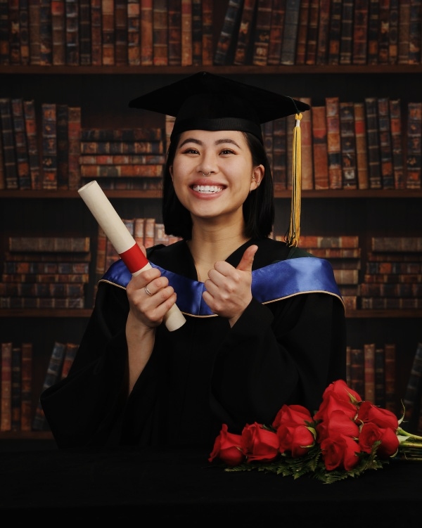 young woman in graduate cloak and hat holding a rolled up parchment in front of a scholarly backdrop