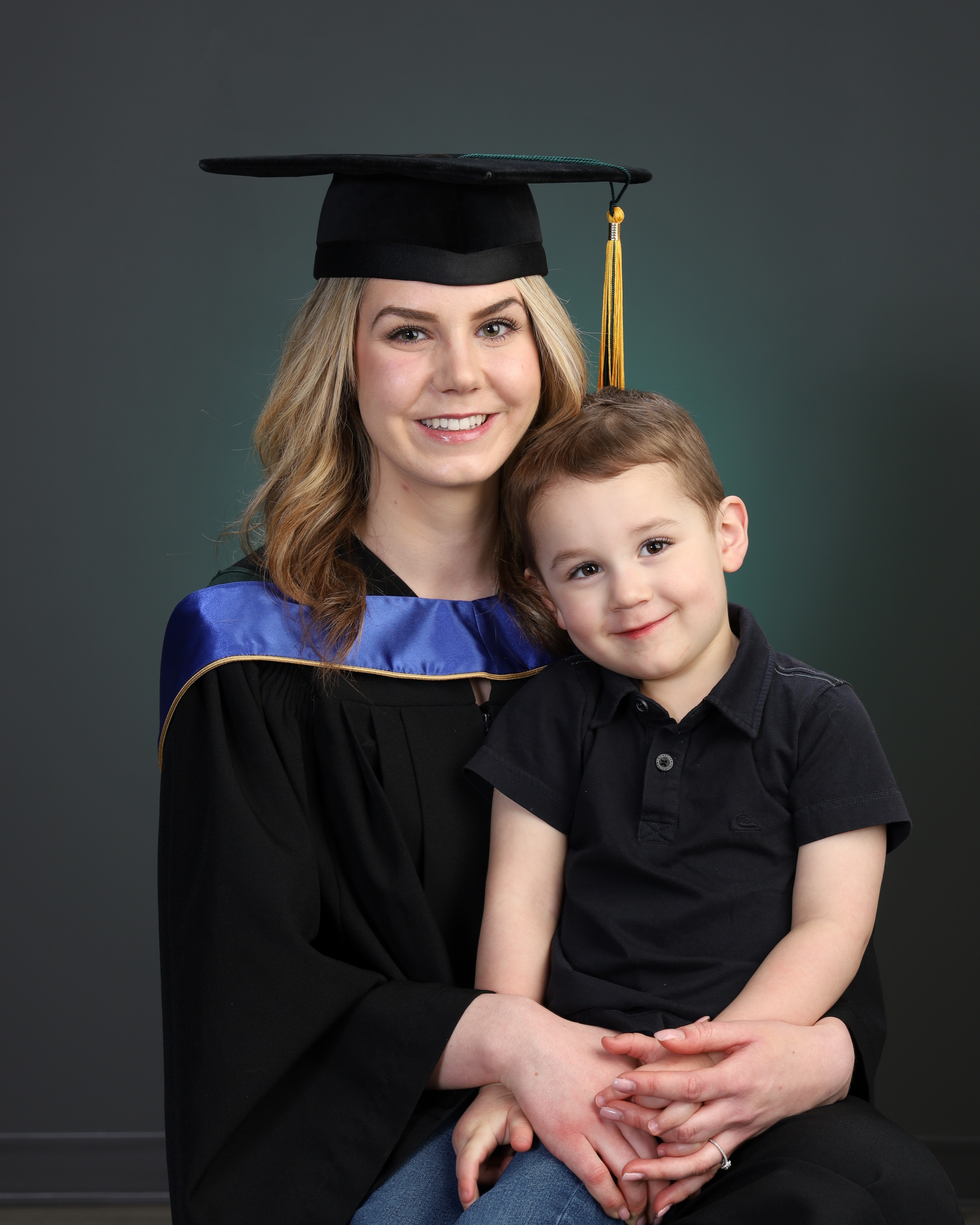blond woman in cap and gown poses for photo with young son in a black shirt on her lap
