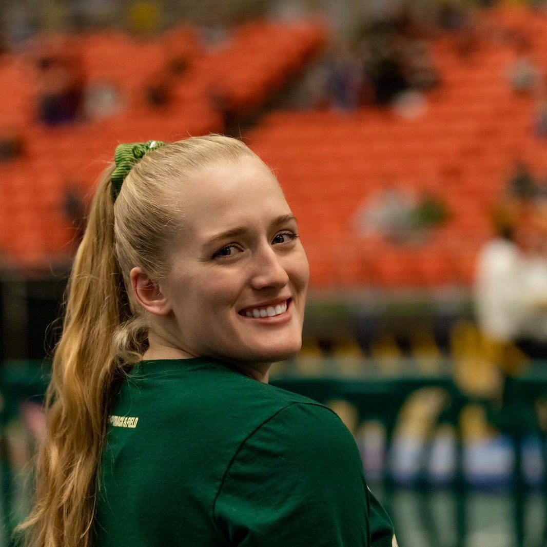 young woman looking over her right shoulder, smiling into the camera in a gymnasium with bleacher blurred out in background