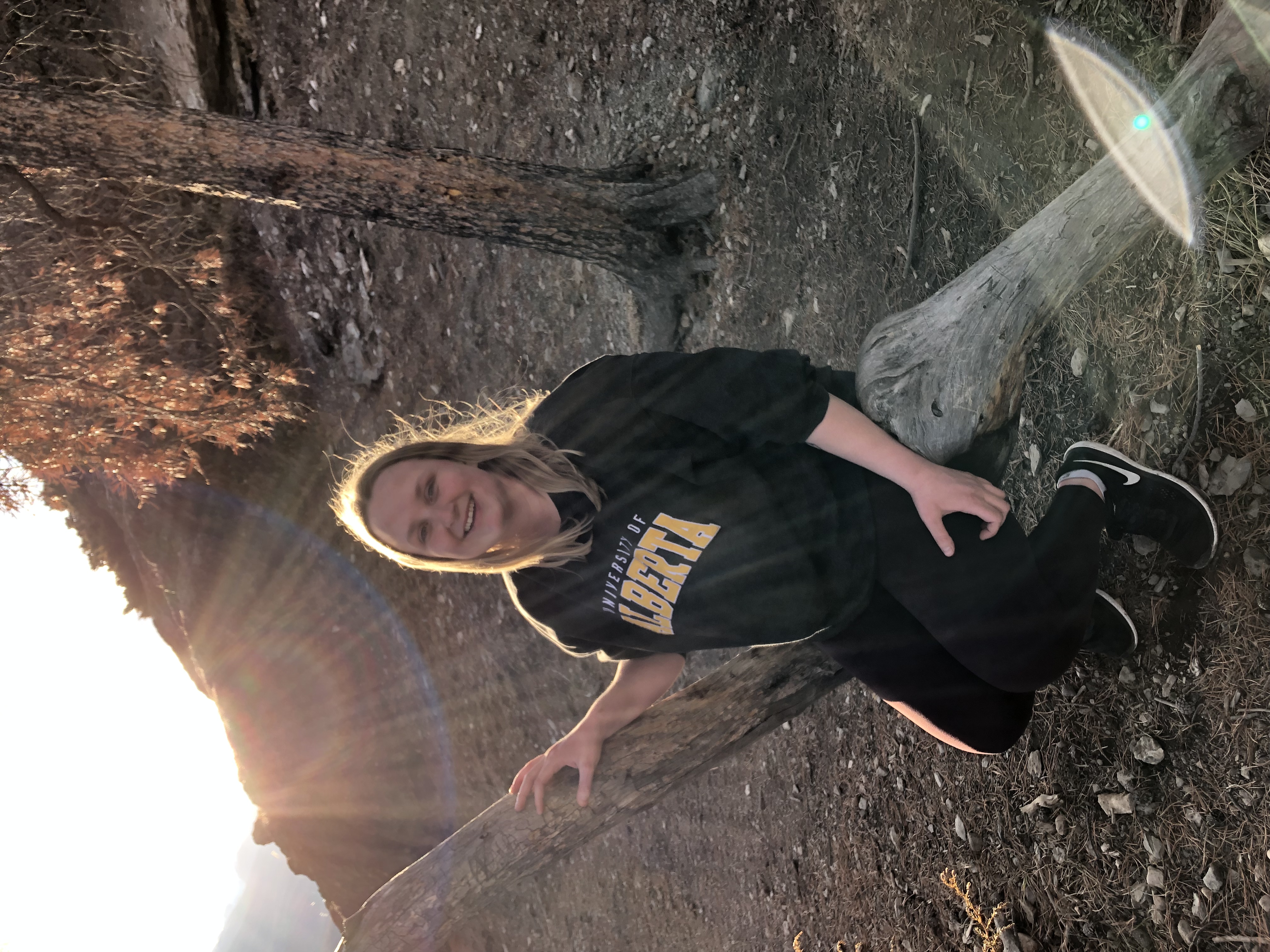 blonde woman wearing a green University of Alberta sweatshirt and black pants sits on tree in wooded area, smiles at camera