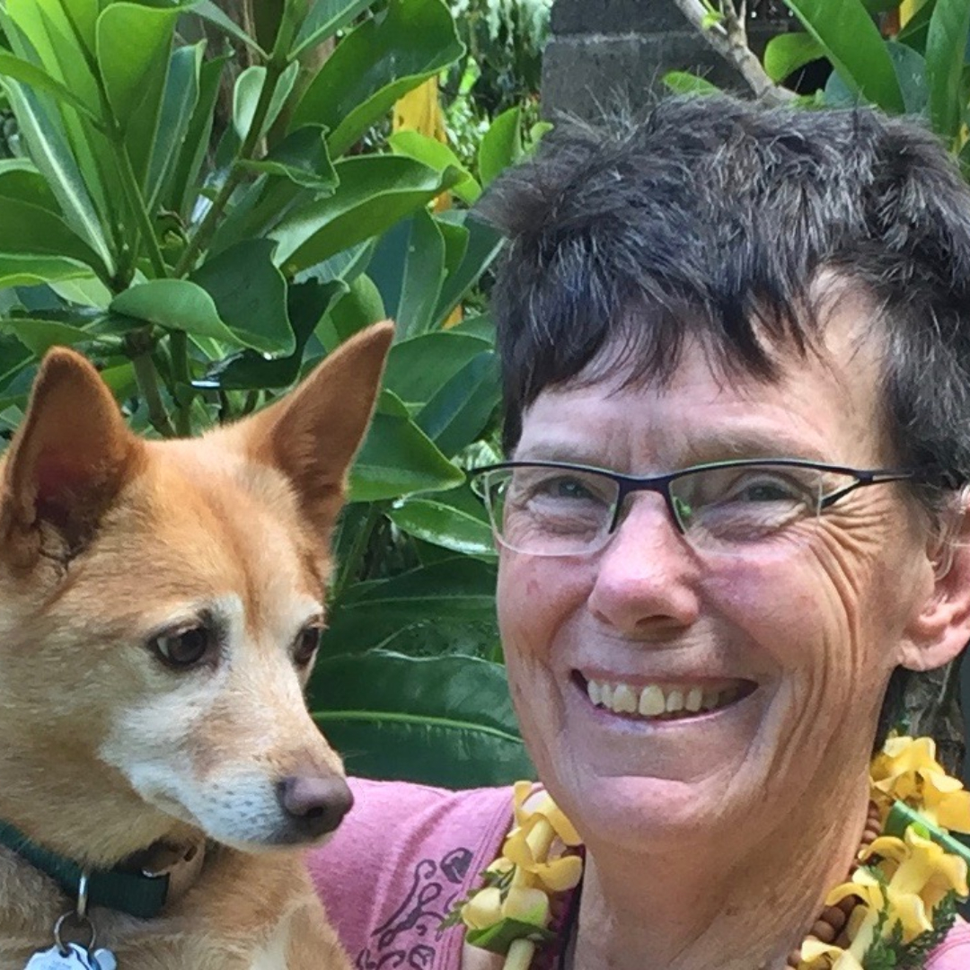 woman in short brunette hair wearing a pink shirt and a yellow Hawaiian lei, holding a golden-coloured dog