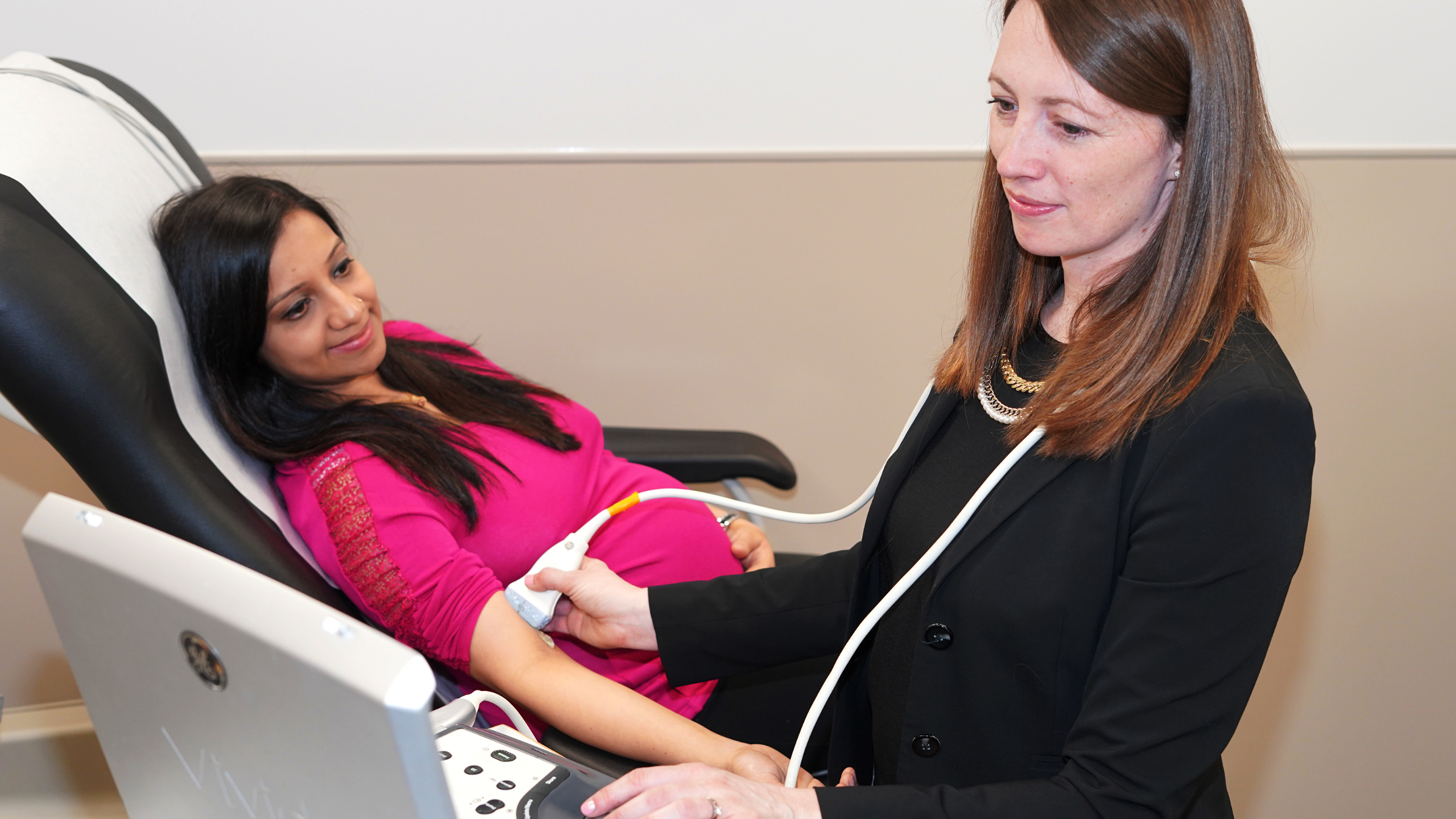 dark-haired woman in a black shirt and black blazer stands in front of patient taking an ultrasound of patient’s arm. Pregnant, dark-haired female patient in dark pink shirt lies back on doctor’s chair while receiving ultrasound on arm