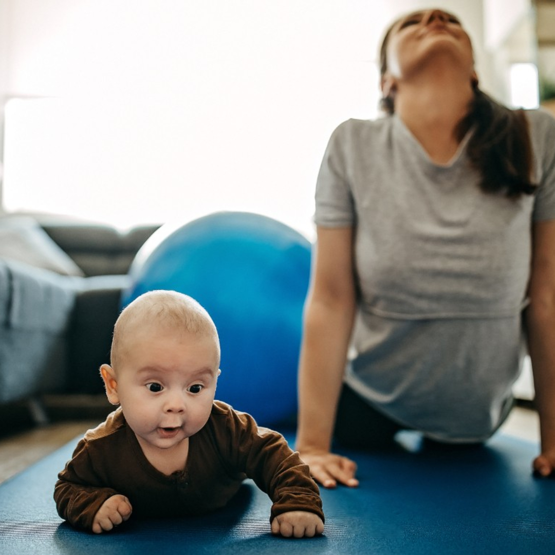 mother in background doing yoga at home with baby crawling in foreground of photo