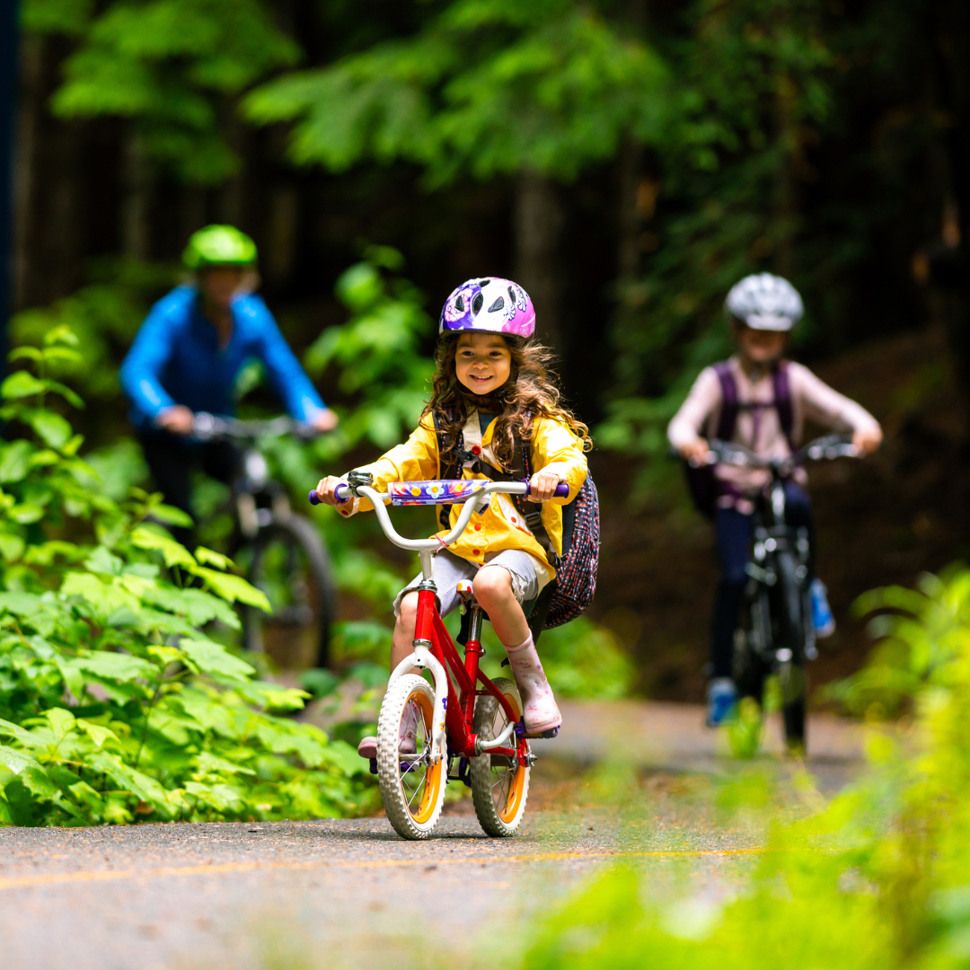 Little girl in yellow rain jacket and pink bike helmet wearing a backpack is riding her pink bike down a trail surrounded by trees, two adults are in the background also on bikes out of focus
