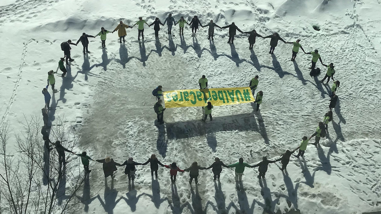 people hold hands in a circle outside in the snow surrounding a banner that reads #UAlbertaCares
