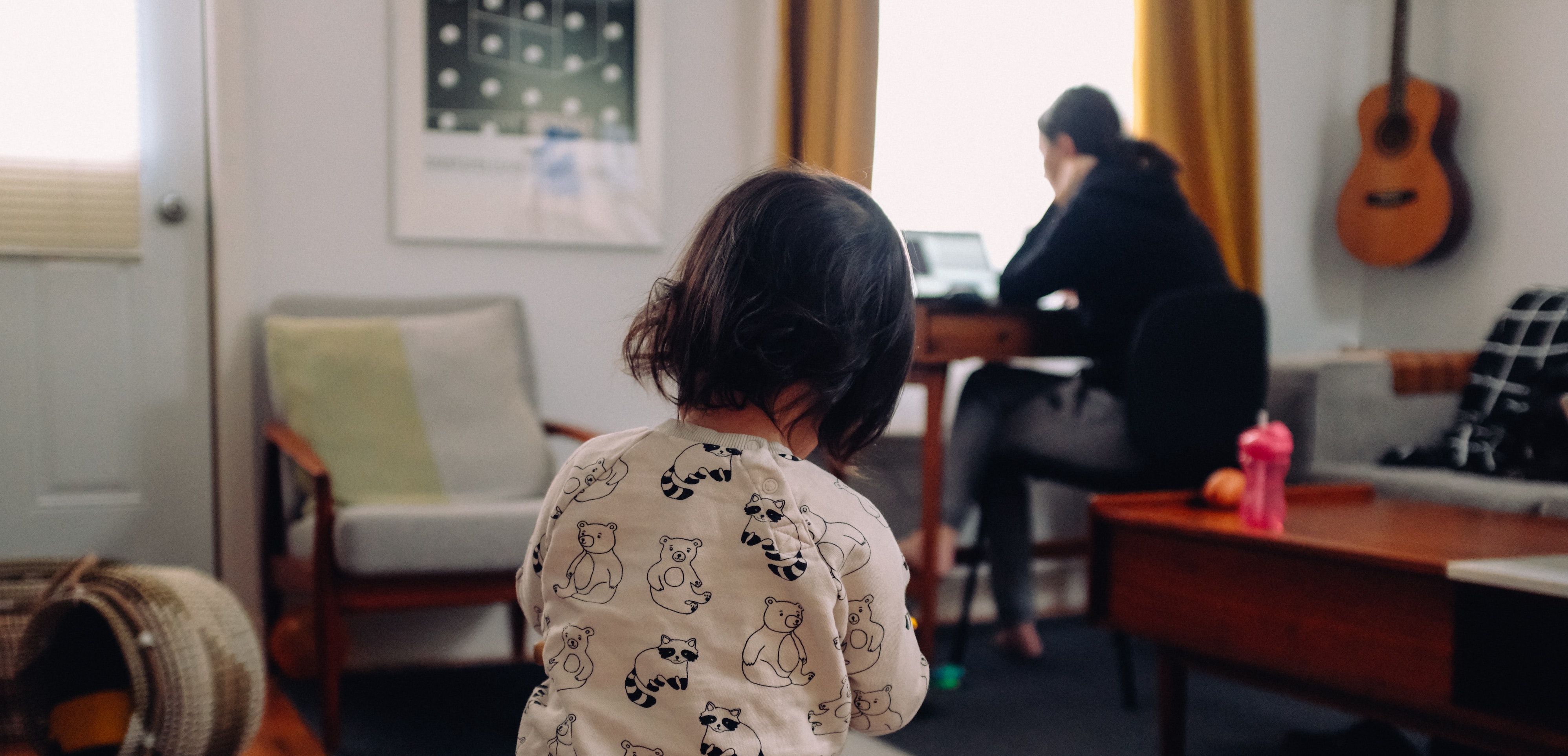 female guardian works on a laptop in the living room while a child wearing bear pajamas plays on the floor