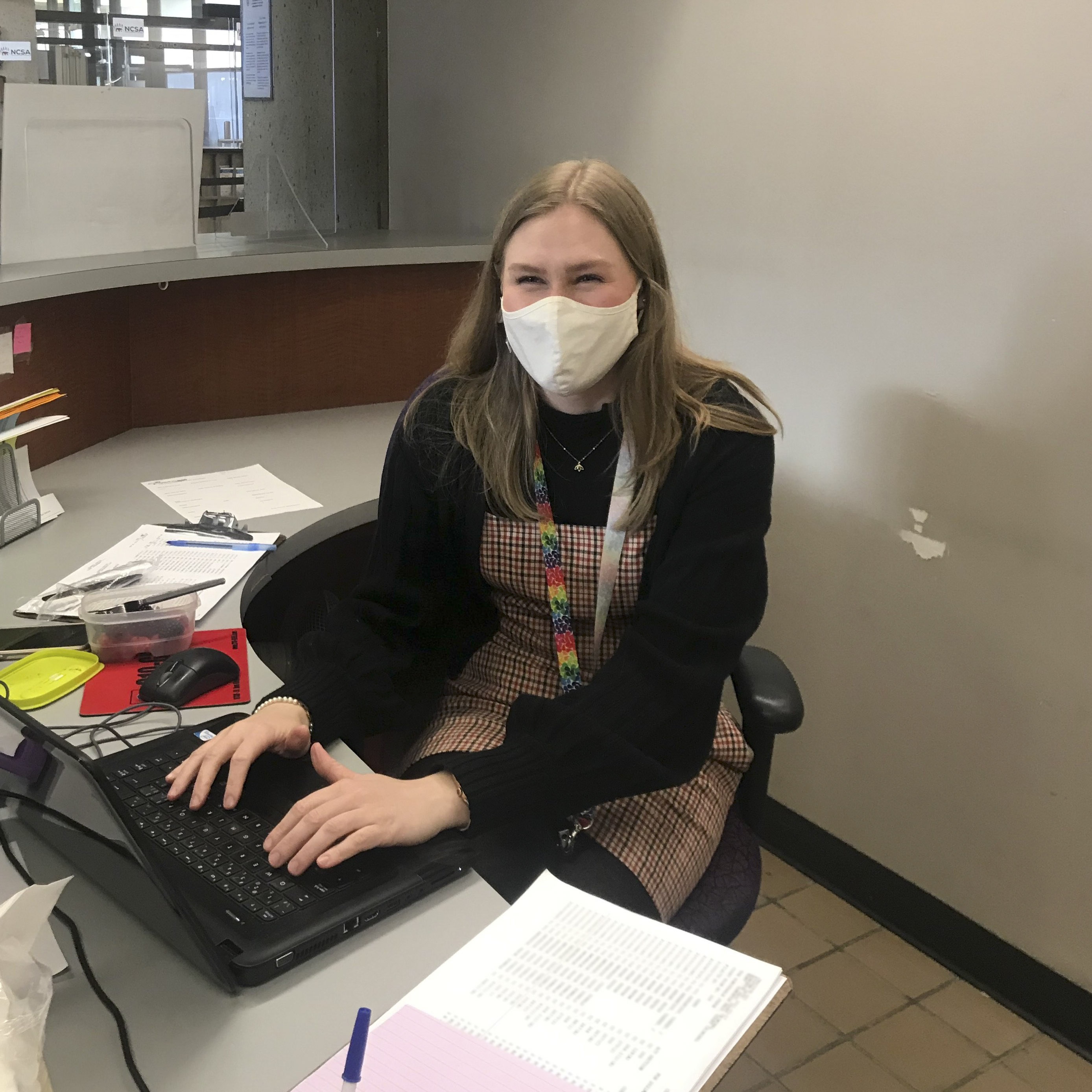 Danielle sits at a desk surrounded by paperwork and is typing on a laptop. She appears to be smiling at the camera even though she's wearing a mask.