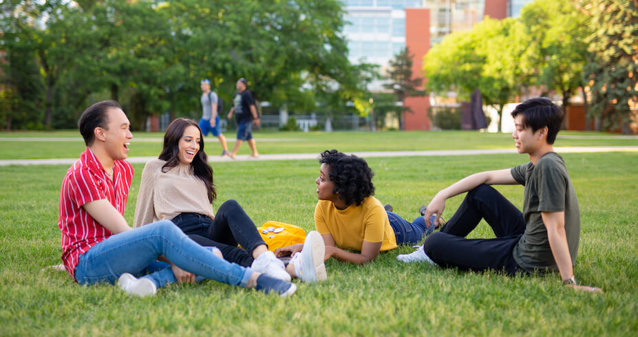Four students sitting and smiling at each other in Main Quad on North Campus.