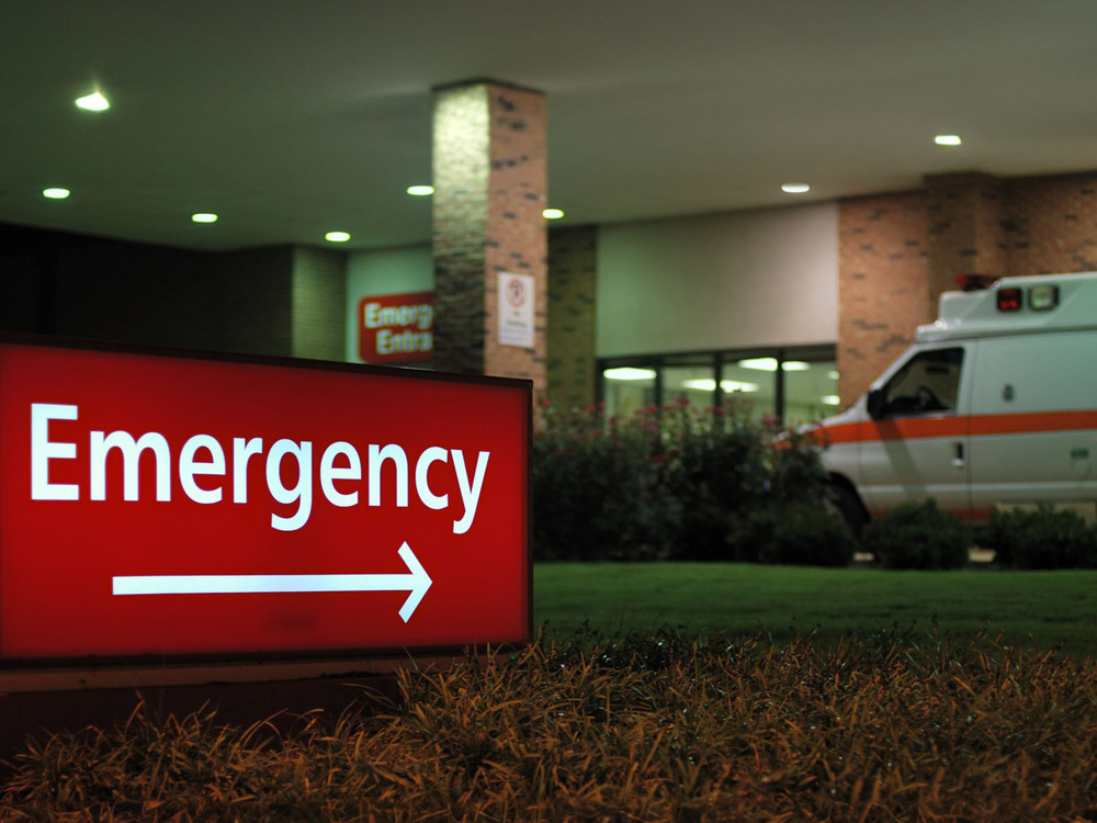 An emergency room sign in dusk lighting with an ambulance and hospital sliding doors in the background.