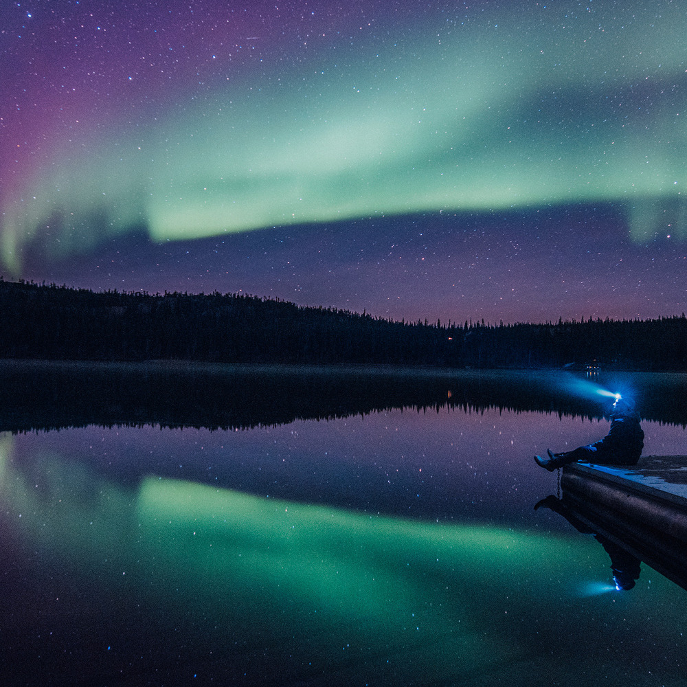An individual looks out at the Northern Lights while seated on a pier at a lake during night.