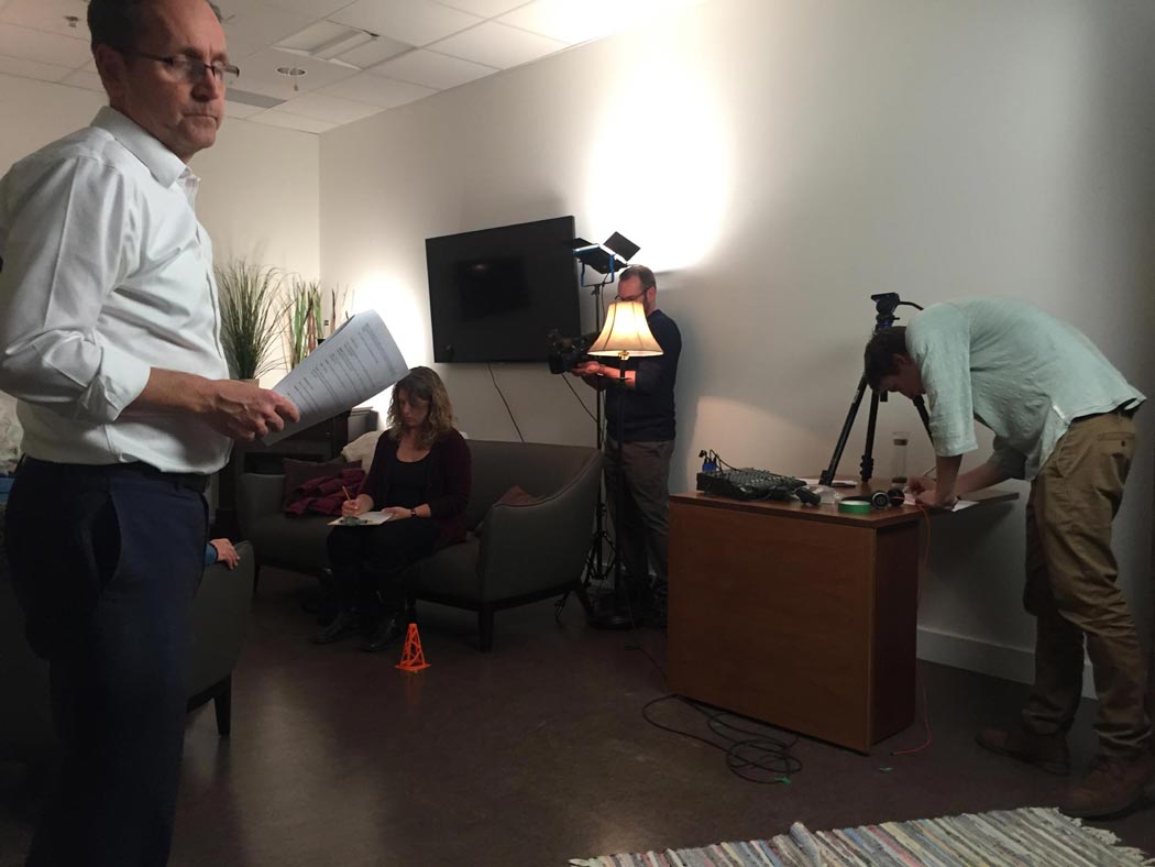 Behind the scenes of the EFS video shoot, a man holds rolled up papers, a woman works on a couch, and a man looks at papers beside a tripod