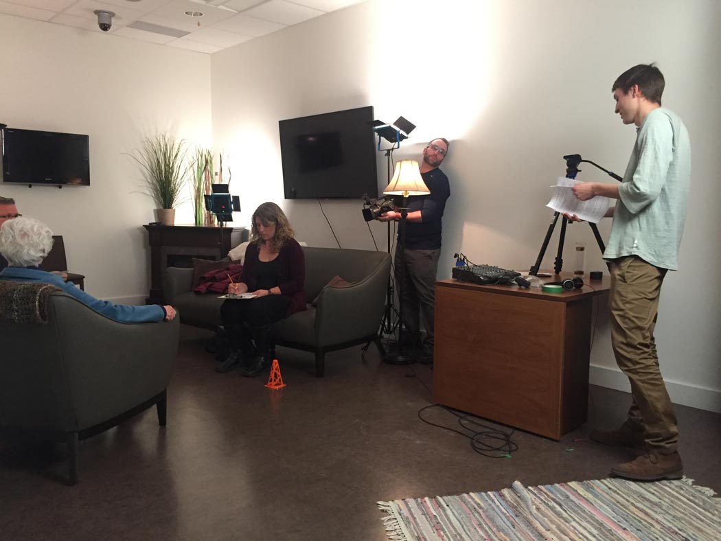 Behind the scenes of the EFS video shoot, a woman works on a couch, a man watches from behind lights, and a man reads from papers beside a tripod